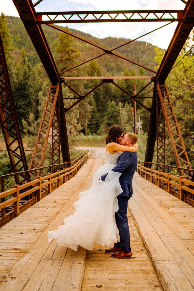 A groom hugs and lifts up his bride as he kisses her on a rustic bridge in the woods at a Montana wedding venue. Photo by Ashley J Photo, a Montana wedding photographer.
