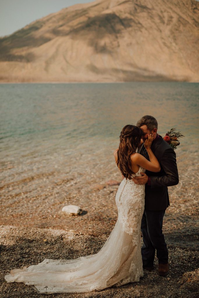 A bride and groom embrace on the waters-edge of a mountain lake in Montana. Photo by Ashley J Photo, a Montana wedding photographer.